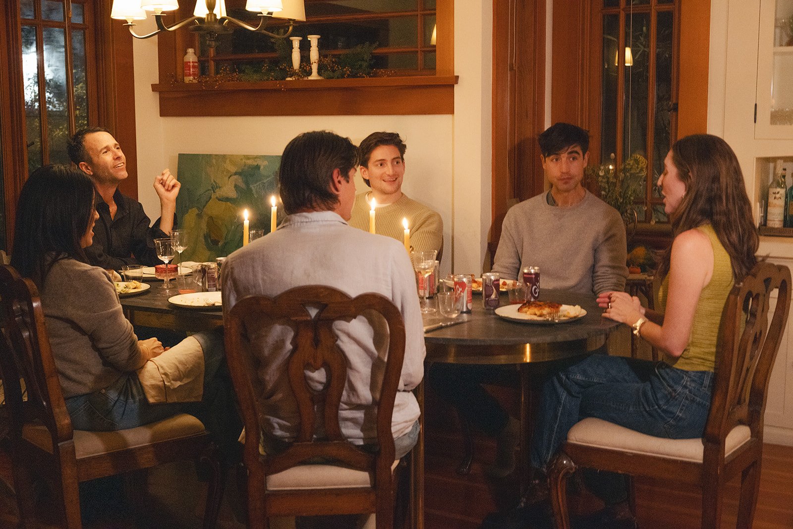 A group of eight people sitting around a dinner table with lit candles, plates of food, and drinks, having a social gathering in a warmly lit dining room.