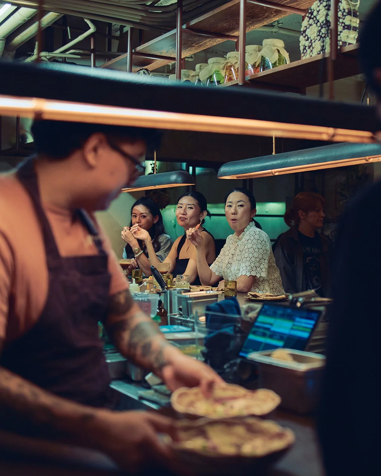 A group of women sitting at a restaurant bar, eating and chatting, with a chef preparing food in the foreground.