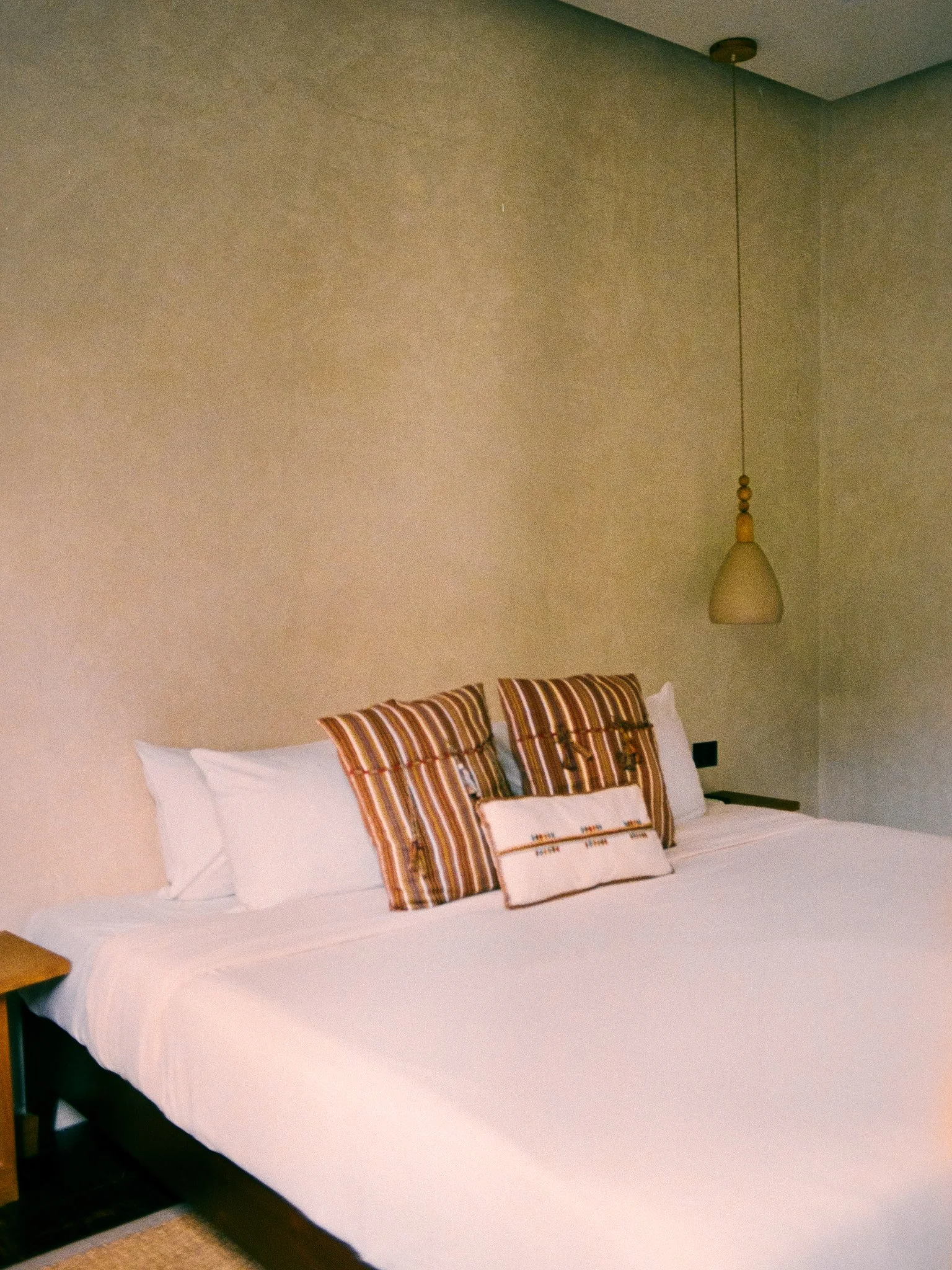 A neatly made bed with white sheets and a white duvet cover, accented by three decorative pillows with striped patterns in brown, orange, and white. The head of the bed is against a plain beige wall, with a wall-mounted lamp hanging above the bed on 