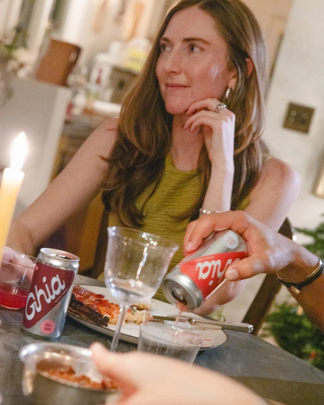 A woman with long brown hair wearing a green sleeveless top sitting at a dinner table, pouring a dry soda into a glass, with a pizza and drinks on the table, in a cozy indoor setting.