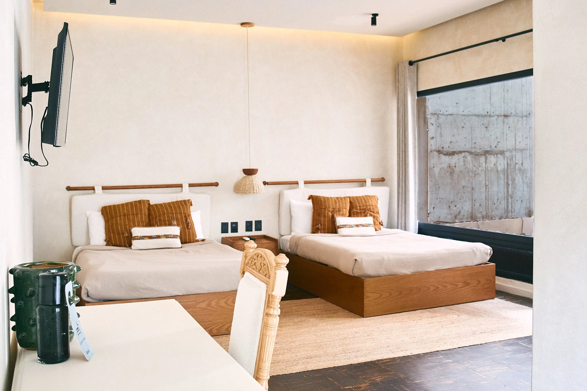 A bedroom with two single beds pushed together, each with white headboards and brown-striped pillows. A small wooden nightstand is between the beds. There is a decorative lamp hanging from the ceiling above the bed area. A sliding glass door with a c