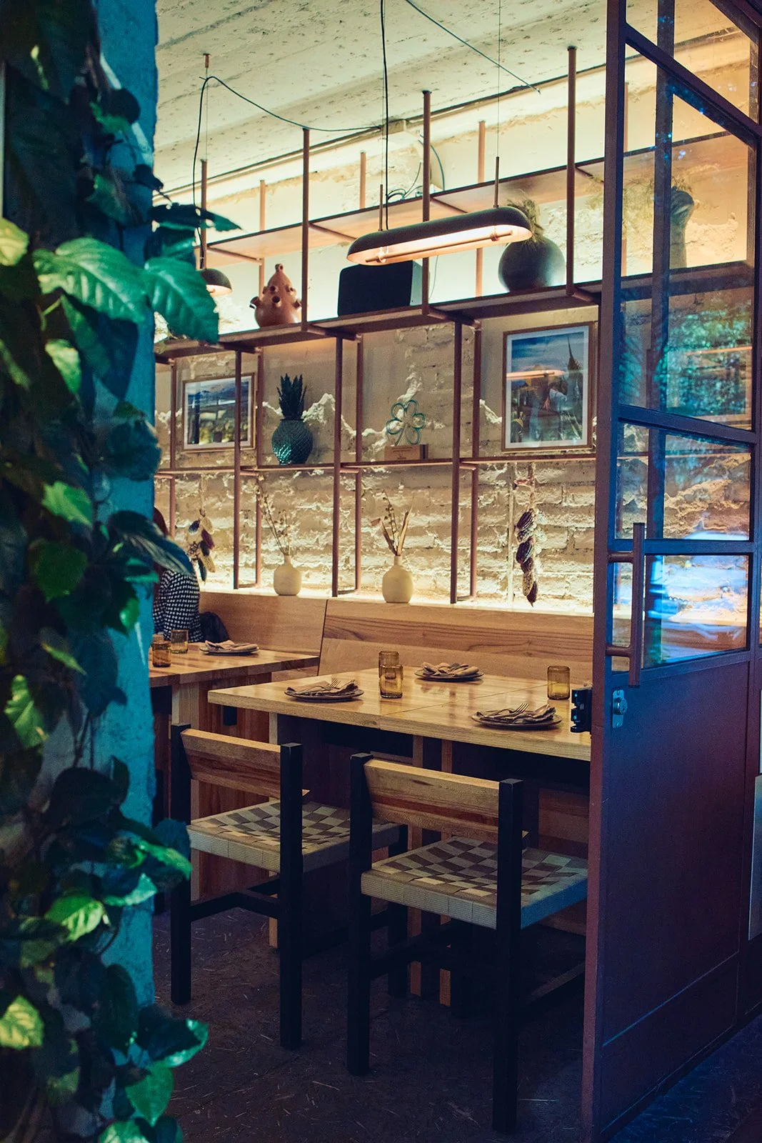 Interior of a cozy restaurant with wooden tables set with napkins, glasses, and plates, decorated with vases and artwork on a white brick wall, illuminated by warm lighting, and partially viewed through an open door with greenery outside.