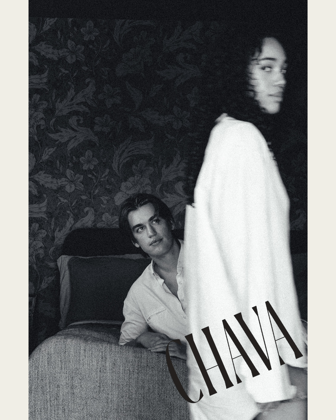 A black and white photo of a woman with curly hair standing in front, and a man with dark hair and light shirt sitting on a bed in the background, with floral wallpaper behind them. The word 'CHAOS' is written diagonally across the woman.