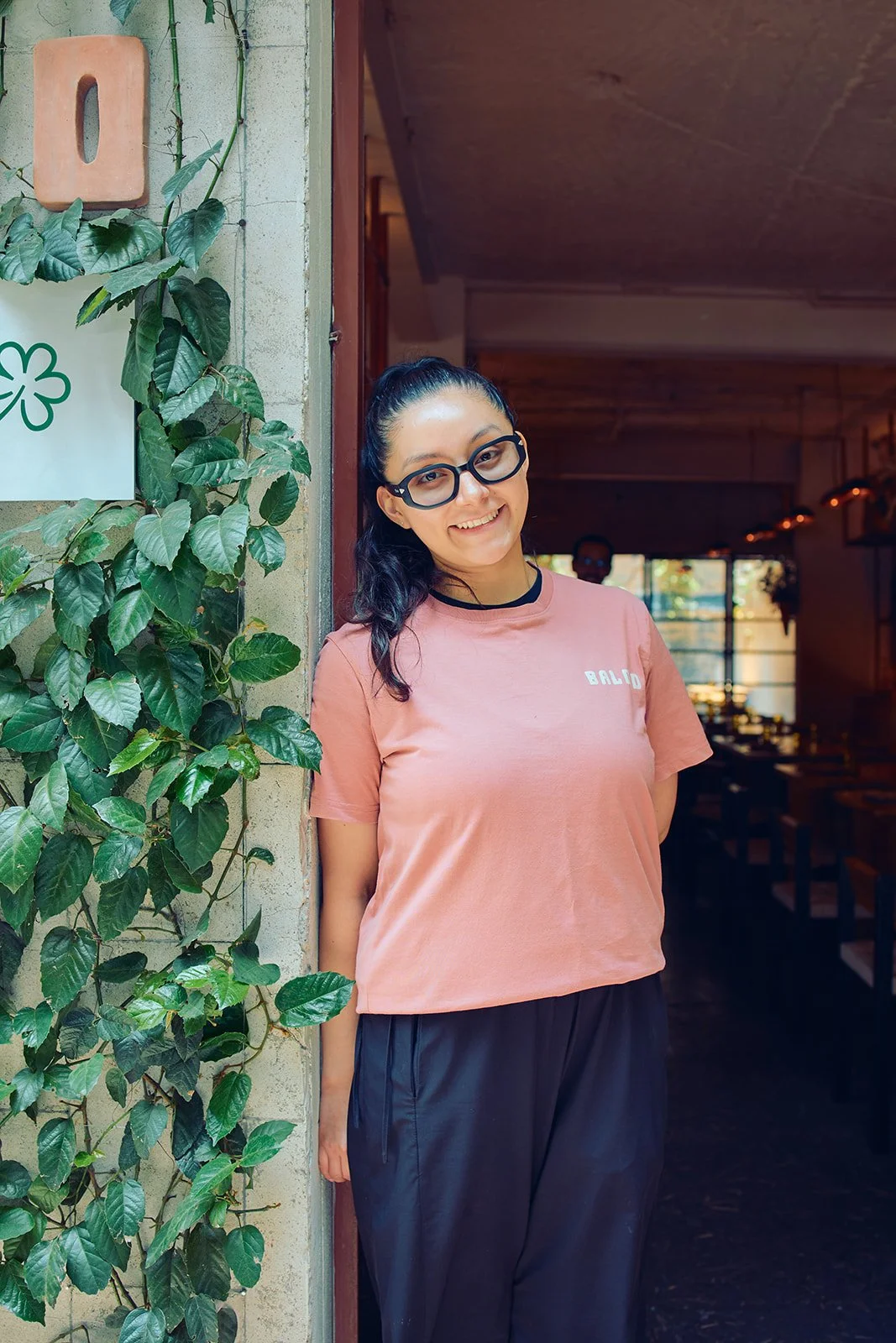 A woman with black hair in a ponytail, wearing glasses, a pink T-shirt with "BALID" on it, and black pants, standing against a wall with green vines, smiling at the camera.