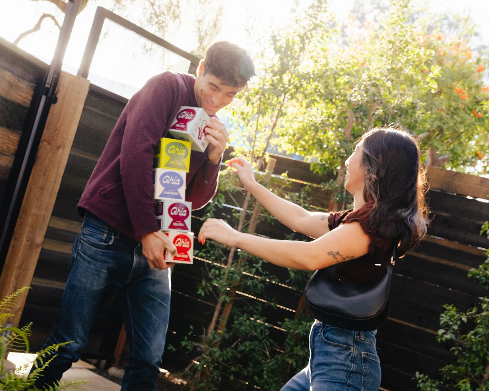 A man and a woman are outdoors, with the woman playfully stacking multiple cartons of Ghia beverage on the man's body. The man is smiling and holding some cartons, while the woman is reaching out to add another carton. They are near a wooden fence and greenery, with sunlight filtering through the trees.