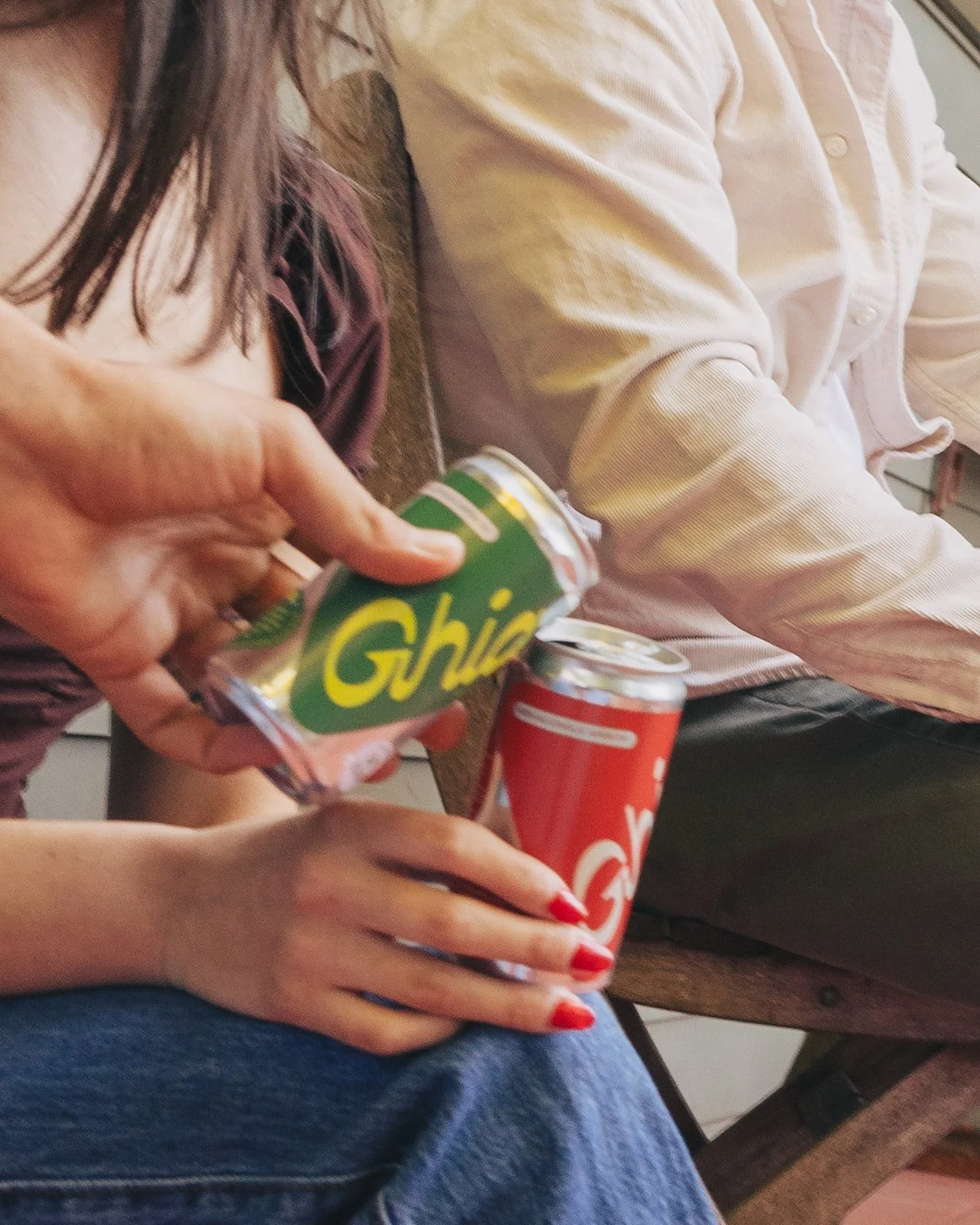 Person with red nails holding a red Coca-Cola can with a hand preparing to open a green Chia can, seated at a table with another person in a white shirt.