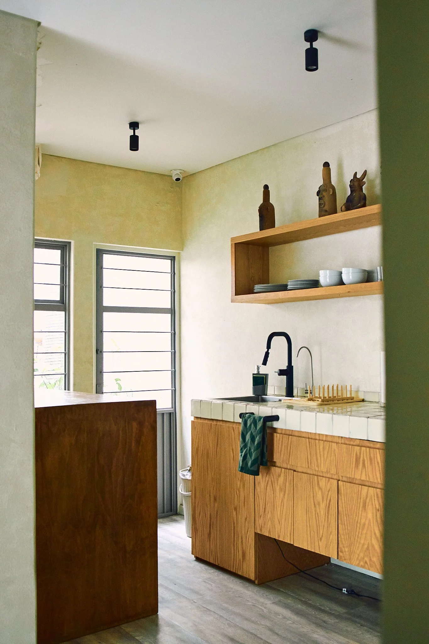 View of a small kitchen with wooden cabinets and a tiled countertop. Open shelf with plates and decorative bottles. Two windows with shutters, a black faucet, and a dish drying rack. Empty ceiling with two black ceiling light fixtures. Light-colored 