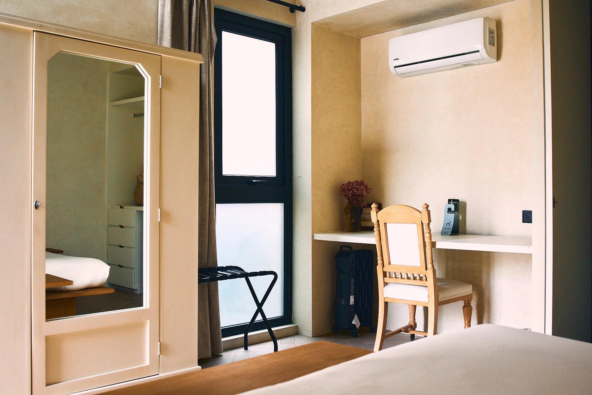 Room with a wardrobe mirror, a window with brown curtains, a white air conditioner, a small desk with a chair, a desk lamp, a potted plant, a black suitcase, and a folding luggage rack.