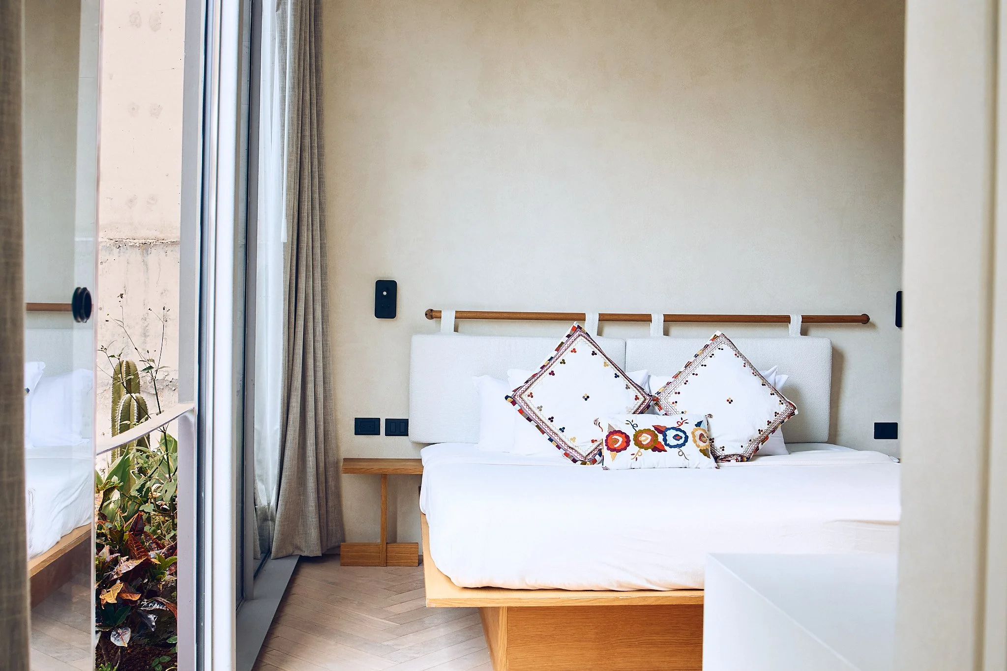 A bedroom with a white bed, colorful pillows, a wooden headboard, and a sliding glass door with beige curtains leading to a balcony with plants.