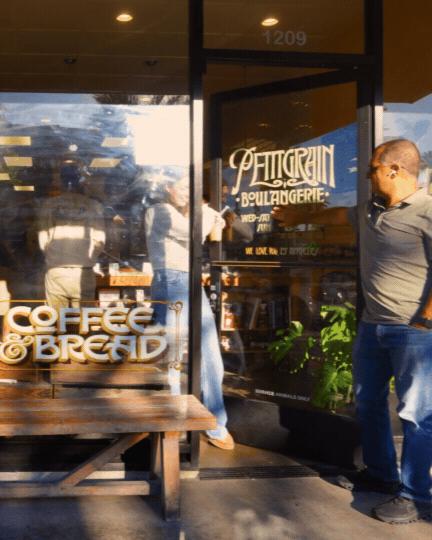 People standing outside a bakery and coffee shop, with a wooden bench in front and large glass windows reflecting sunlight.