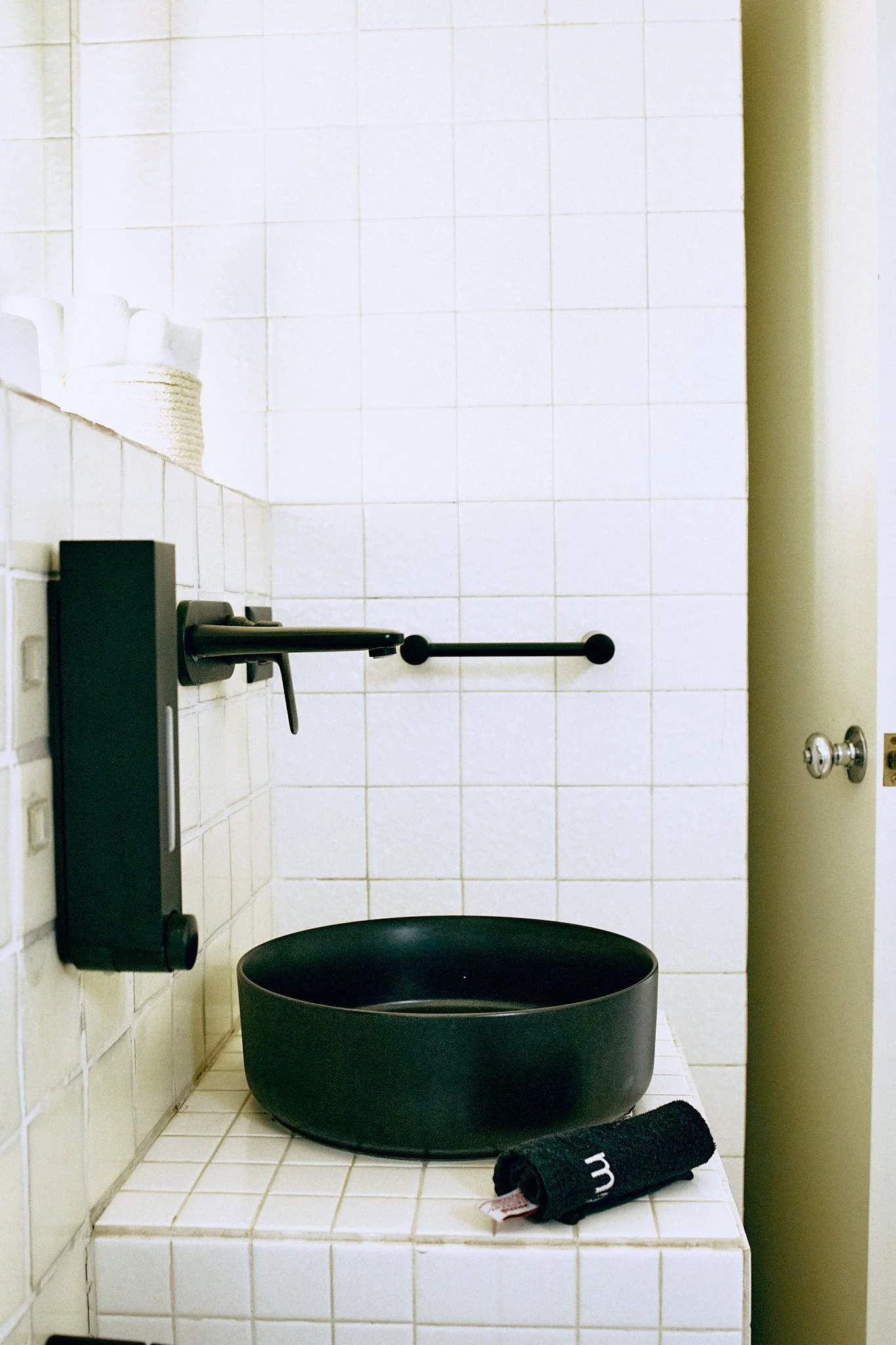A bathroom with white tiled walls and floor, black rectangular bathtub, black towel rolled on the tiled ledge, a black grab bar on the wall, and black fixtures.