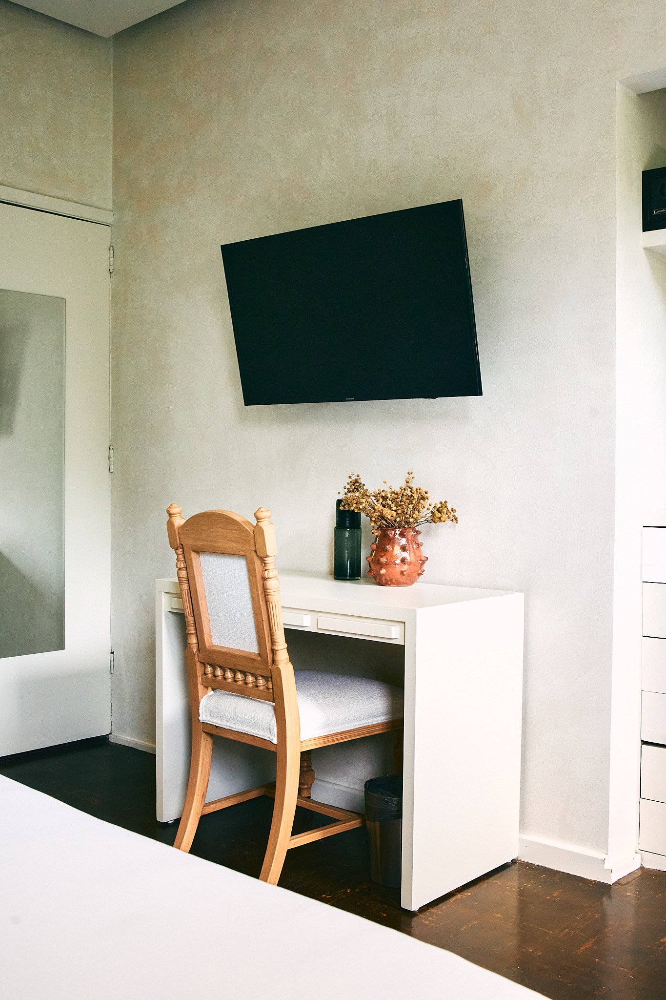 A small white desk with a wooden chair in front of it, decorated with a pink vase of dried flowers, a green thermos, and a small black container, situated against a beige wall with a wall-mounted flat-screen TV above.