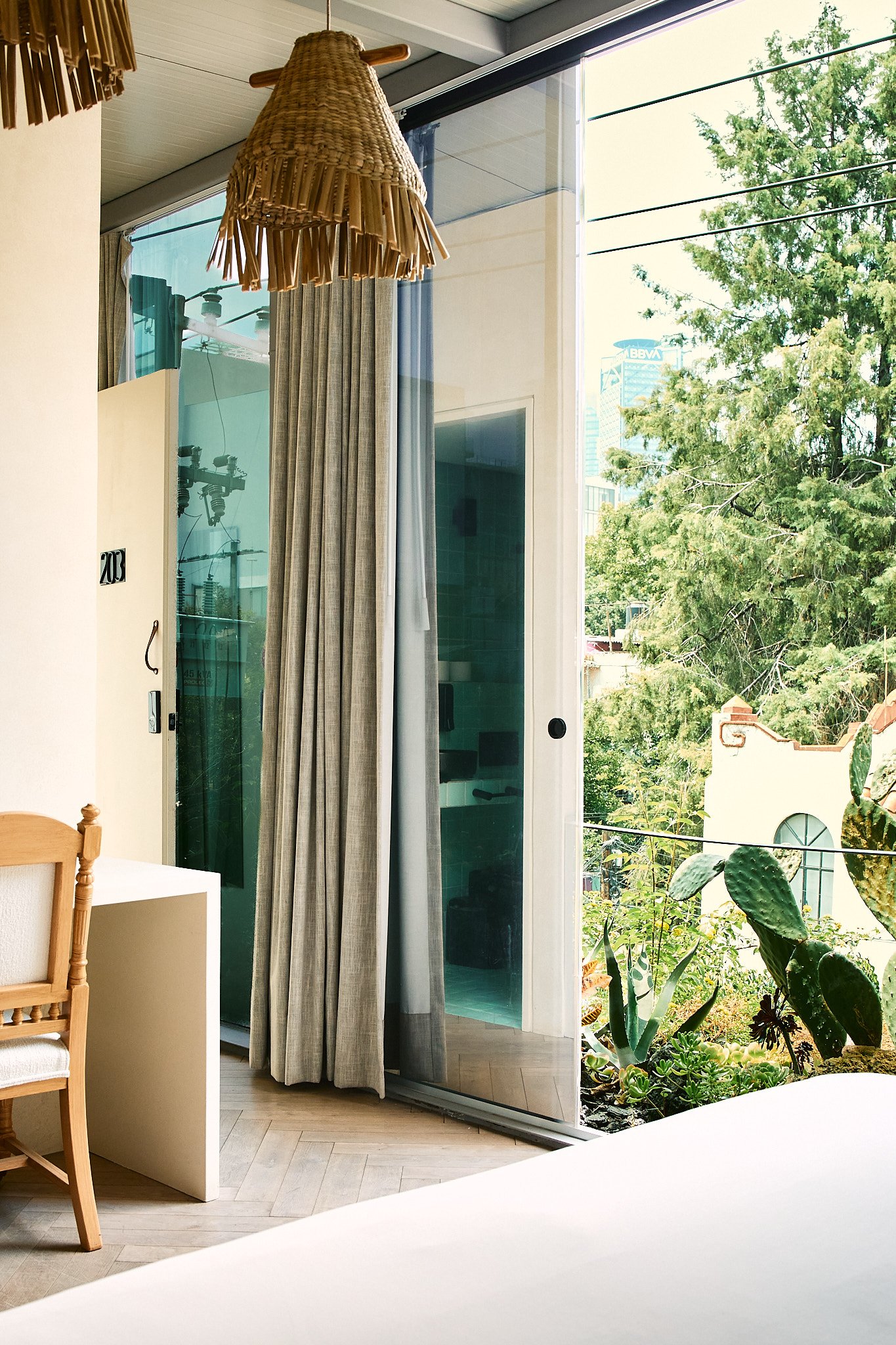 View of a room with a large glass sliding door opening to a garden with greenery and trees, a cactus, and a partial view of a building with a tower in the background, a woven hanging lampshade, and beige curtains.