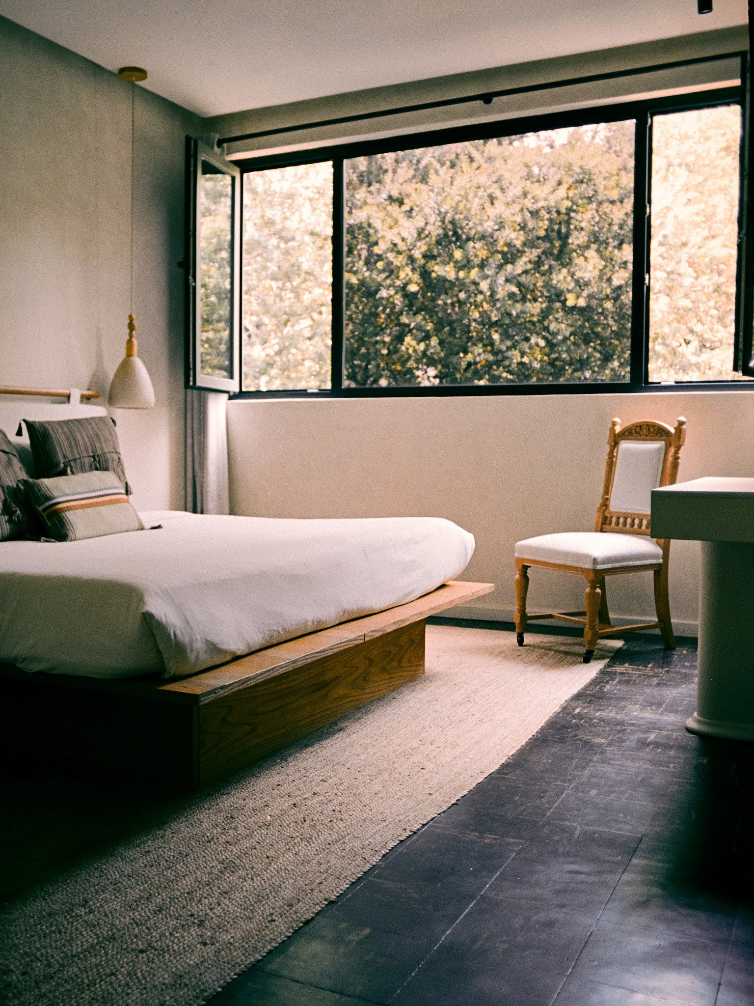 A minimalist bedroom with a large open window showing trees outside. The room contains a low wooden bed with white bedding and plaid pillows, a small pendant lamp hanging beside the bed, a vintage wooden chair, and a side table. There is a large rug 