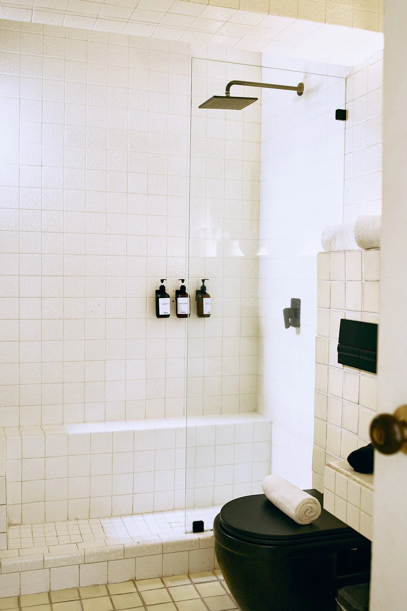 A white tiled bathroom with a walk-in shower featuring a rainfall showerhead. There are three bottles of toiletries on a shelf inside the shower. Below the shower, there is a curved black toilet with a rolled towel on top. To the right of the toilet,
