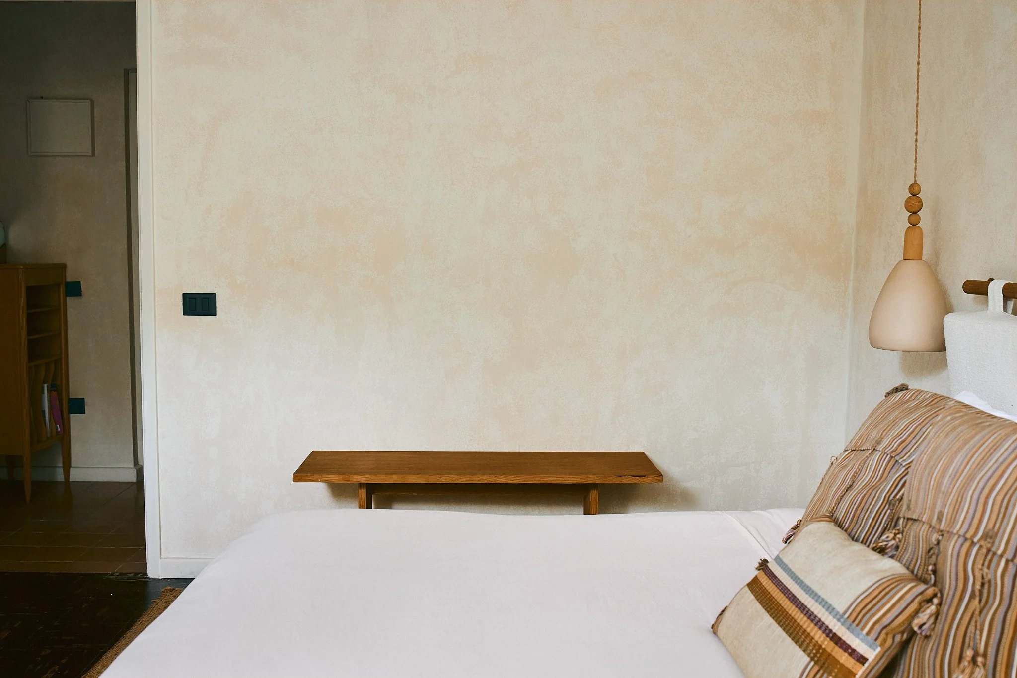 View of a bedroom with a bed, striped pillows, a beige wall, a hanging bedside lamp, a wooden table, and partially visible bookshelf and doorway.