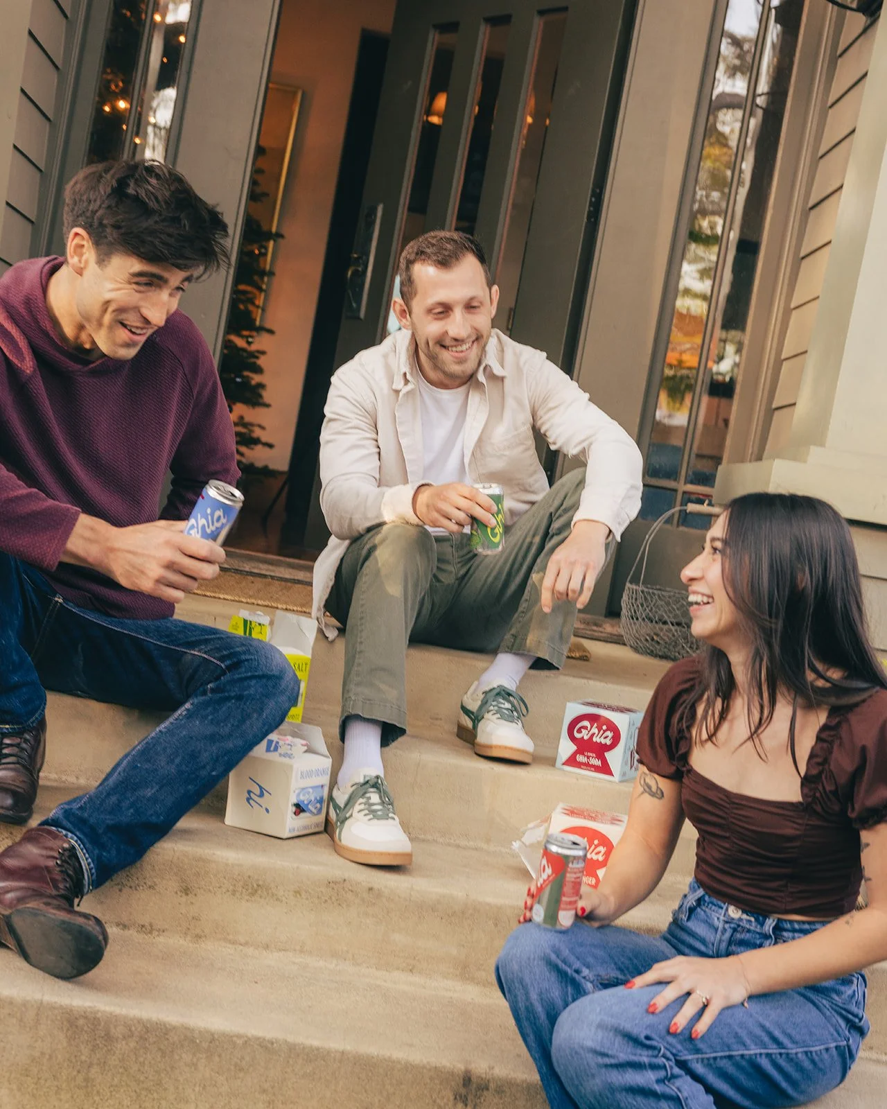 Three young adults sitting on steps outside, smiling and holding cans of soda, with snack boxes nearby, enjoying a casual conversation.
