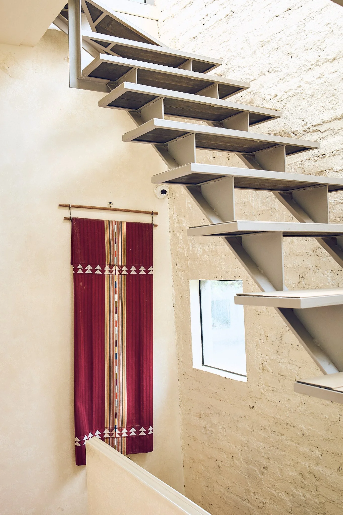 Interior view of a stairway with wooden steps and white railing, beige walls, a small window, and a colorful, striped wall hanging.