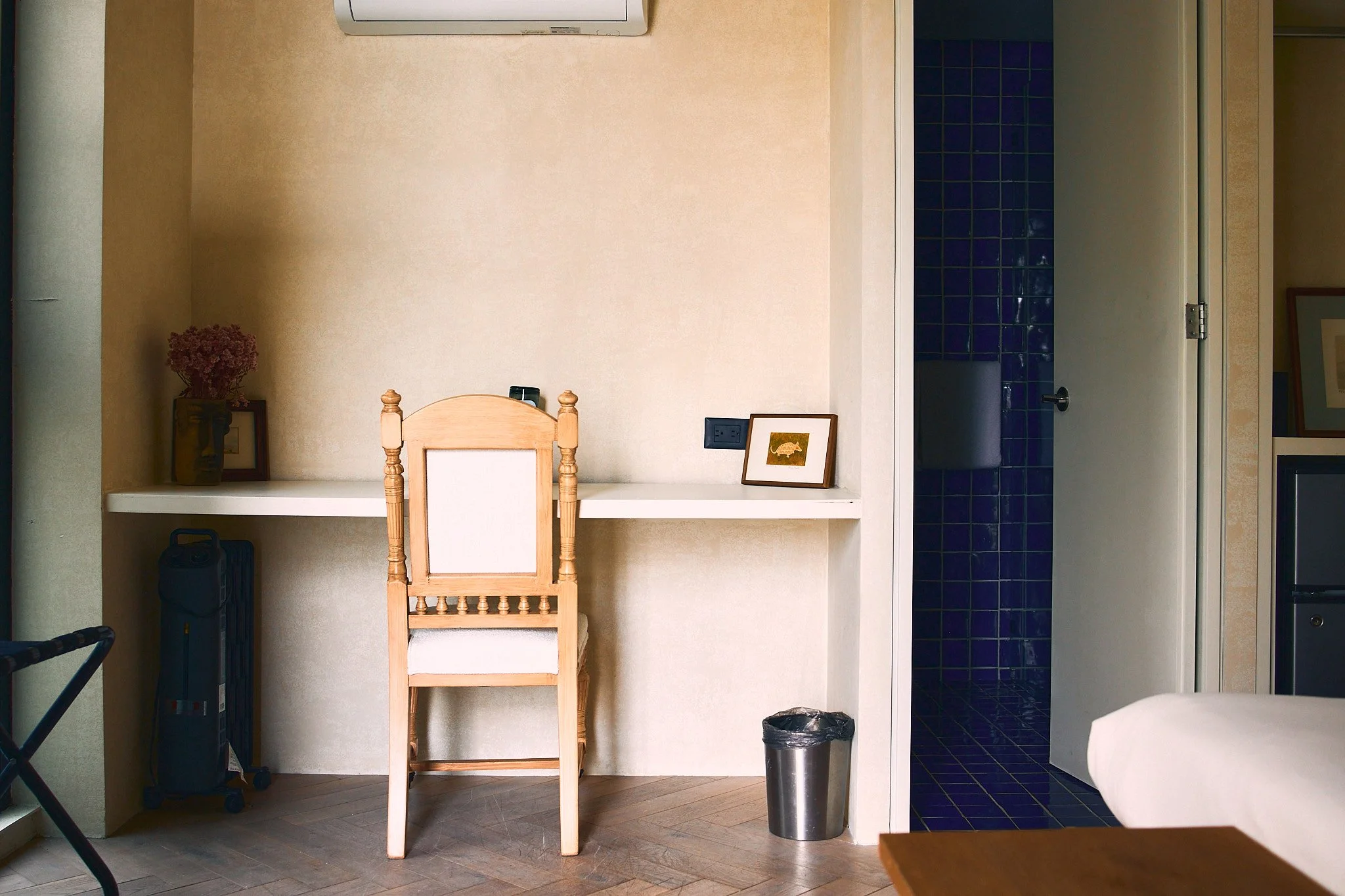 A minimalist interior space with a wooden chair, framed artwork, a vase with flowers, an electrical outlet, and a small trash can, next to a wall and an open door revealing blue tiled walls of a bathroom or kitchen area.