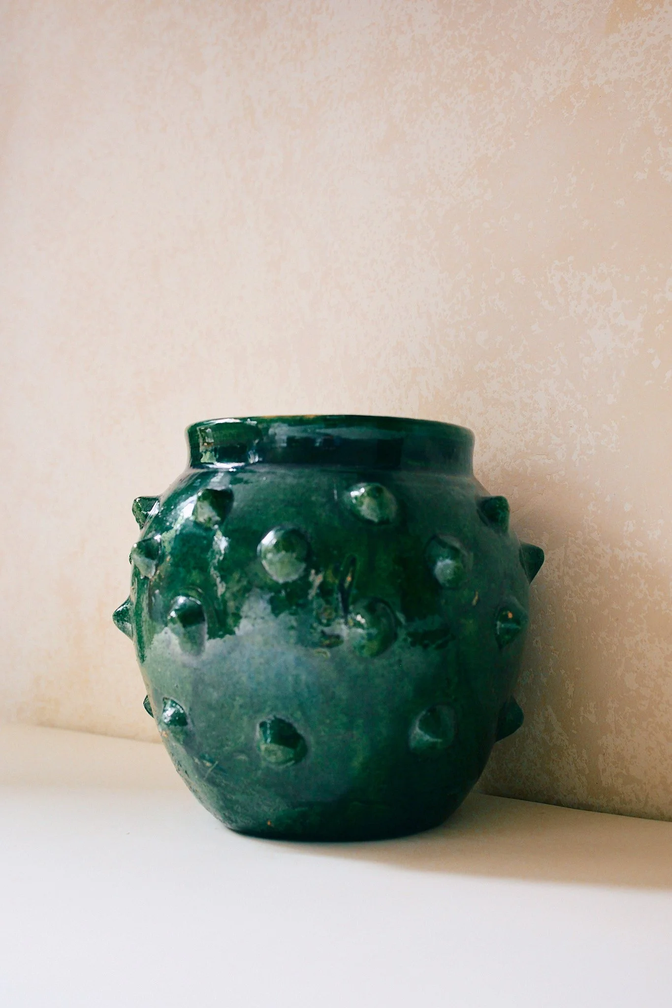 A round green ceramic vase with raised dot patterns on a white surface, against a beige wall background.