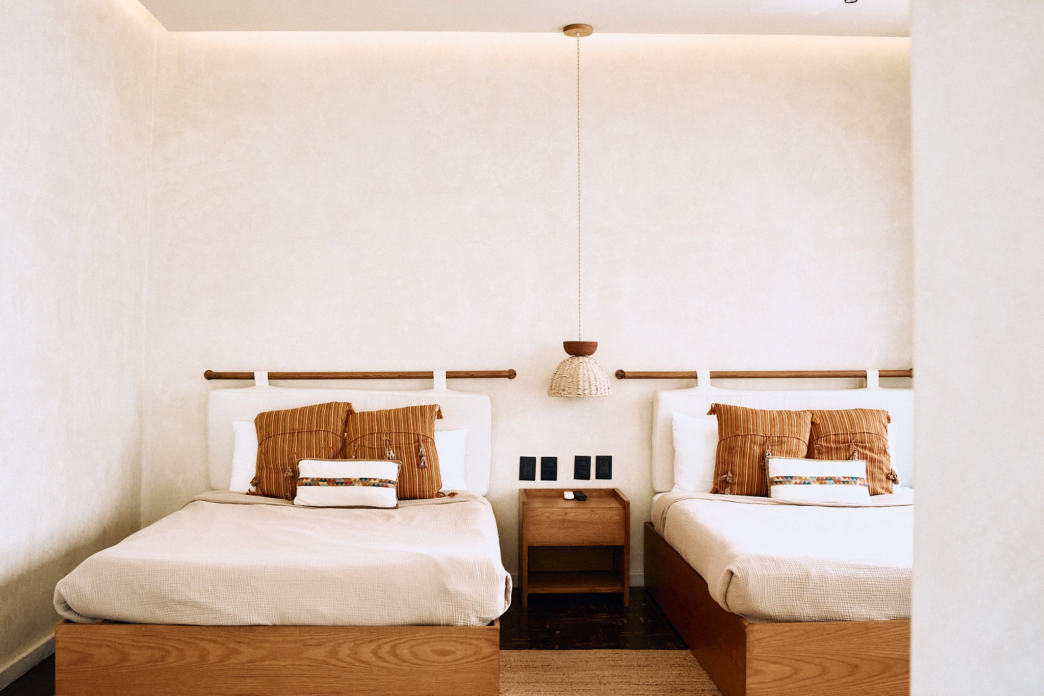 Two beds with white bedding and striped pillows in a minimalist bedroom with soft lighting, a hanging lamp, and a wooden nightstand between the beds.