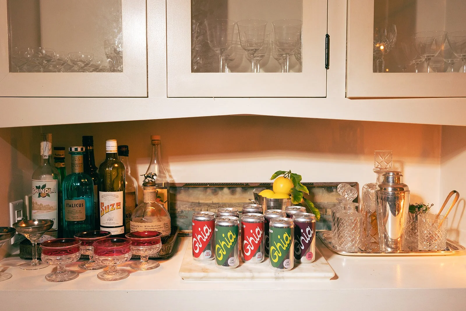 A home bar with various bottles of alcohol, glassware, and canned drinks, including 6 cans of Ghia soda, in a kitchen setting.