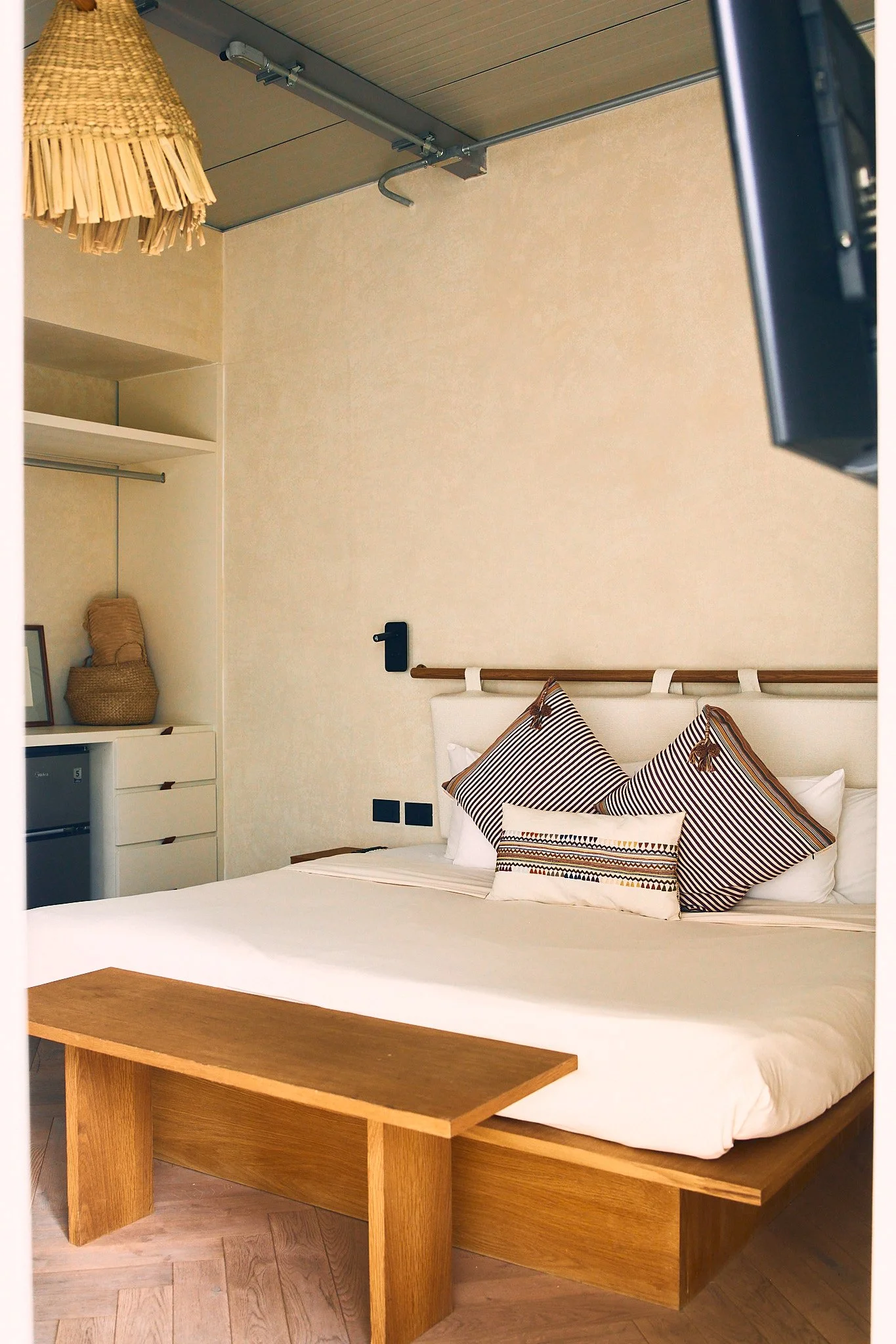 A bedroom with a white bed, decorative pillows, a wooden headboard, and a wooden table at the foot of the bed. The room has beige walls and a built-in shelf with decor and a basket. A black wall-mounted lamp is next to the bed, and a wooden ceiling w