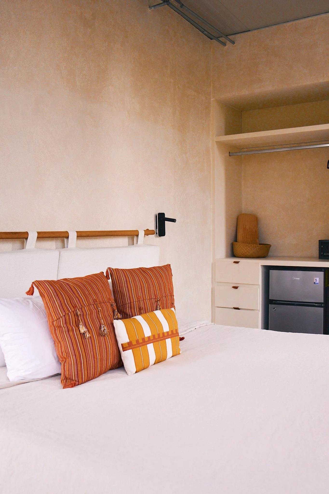 A bed with white bedding and four pillows with orange, yellow, and brown striped covers in a room with beige walls. In the corner, there is a built-in shelf with a small basket and a television.