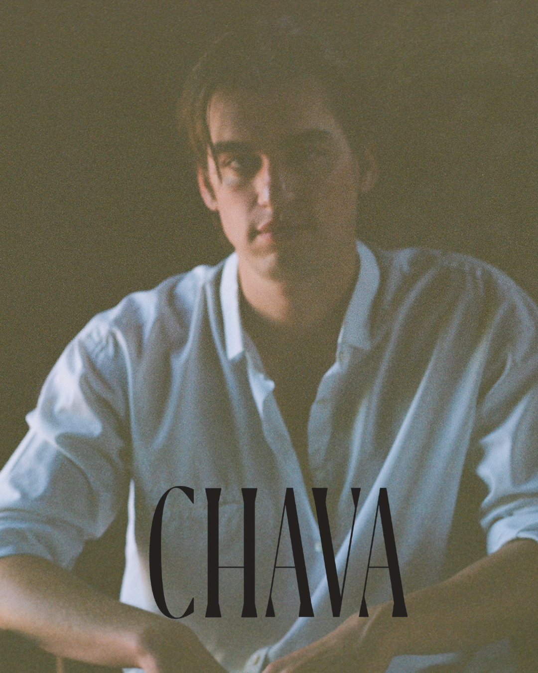 A young man with dark hair wearing a light blue shirt, looking at the camera with a serious expression, in dim lighting with the word 'CHAVA' written at the bottom.