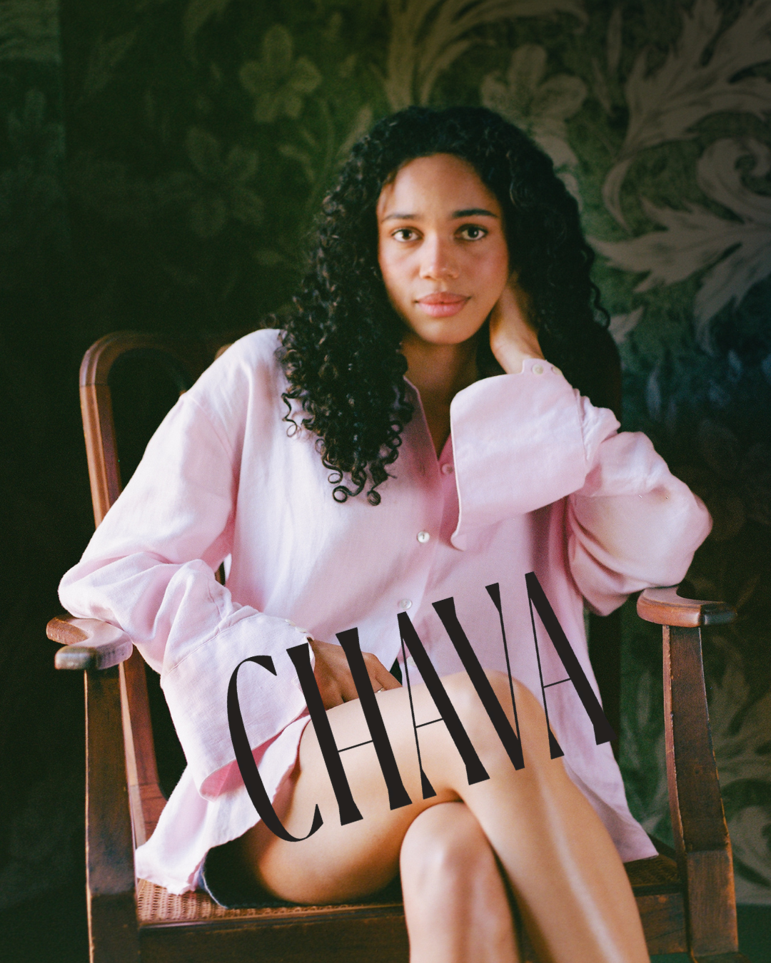 A woman with curly hair wearing a pink shirt sitting on a wooden chair with one hand behind her neck. The word 'CHAIR' is written across her lap. Dark greenery background.