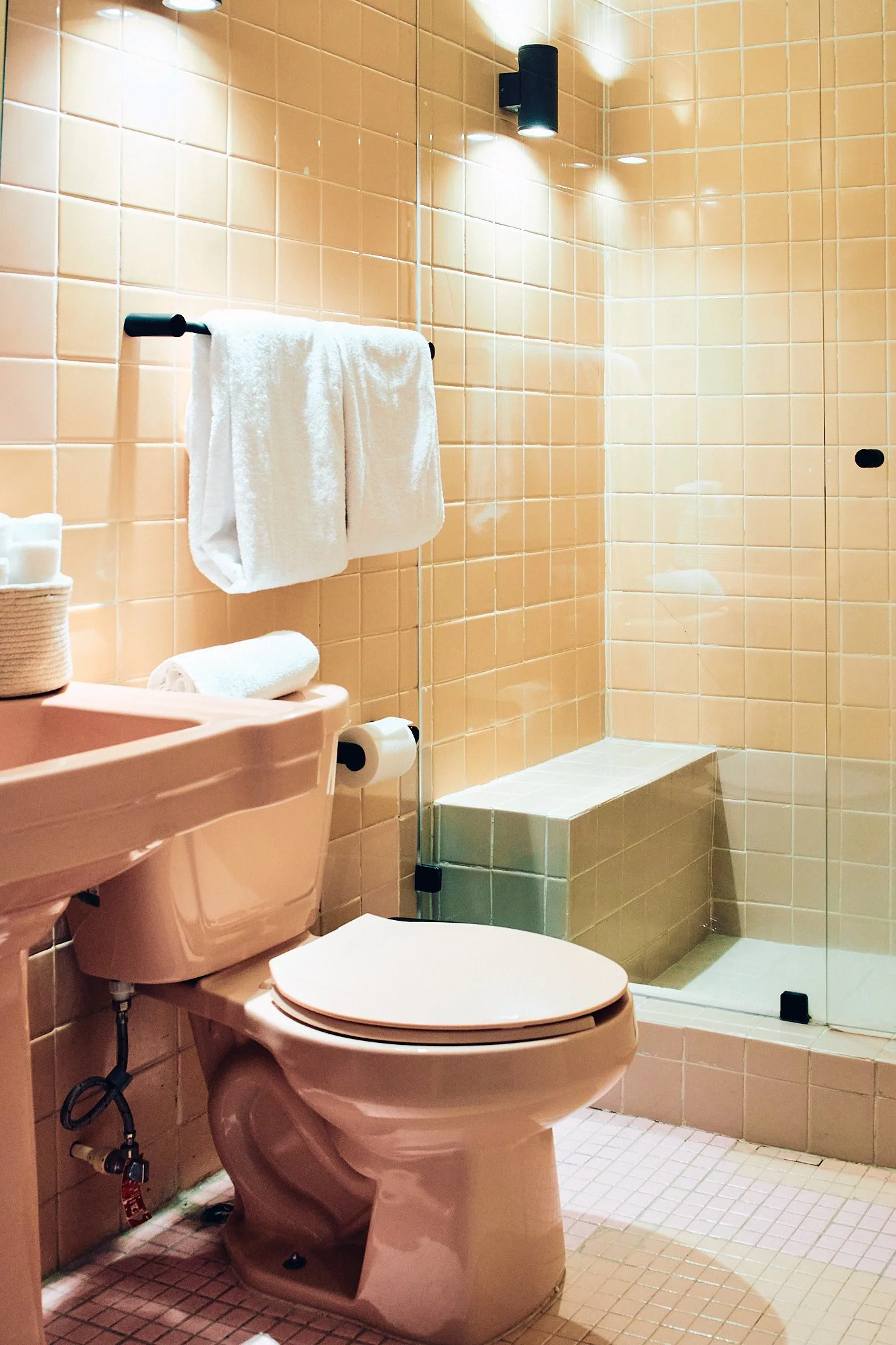 A bathroom with beige tiled walls and floor featuring a pink toilet, a pink sink, white towels, and a glass shower enclosure with beige tiles inside.