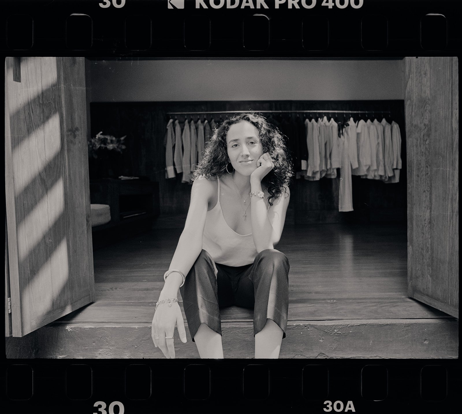 A black and white photo of a woman sitting on a step in front of a closet with hanging clothes. She has curly hair, wears a sleeveless top, hoop earrings, a cross necklace, and bracelets, and is resting her chin on her right hand.