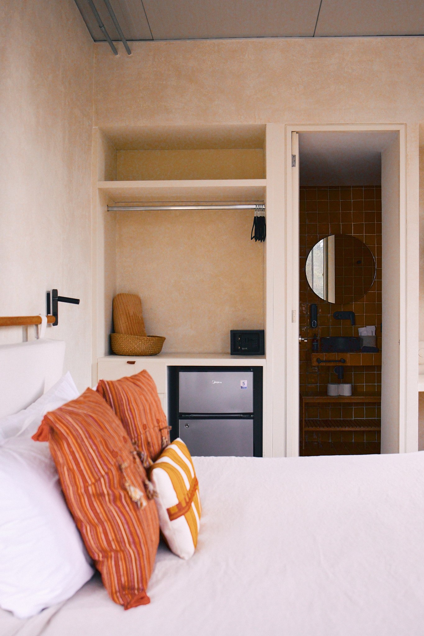 Hotel room with bed, orange striped pillows, a small refrigerator, and a view into a bathroom with brown tiles and a mirror.
