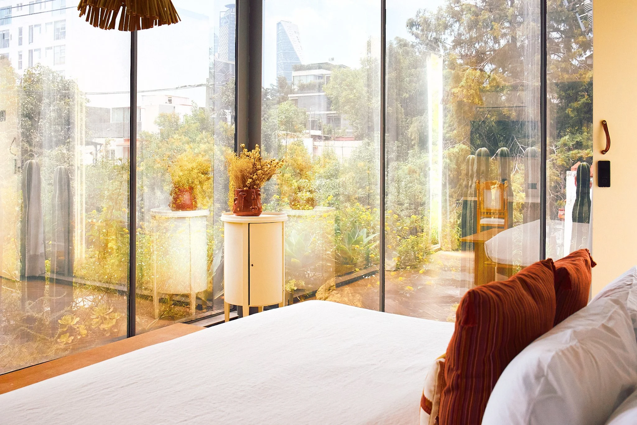 A bedroom with a large glass window showing trees and city buildings outside, decorated with potted plants on a white table, a bed with white bedding and orange pillows, and a brown headboard.