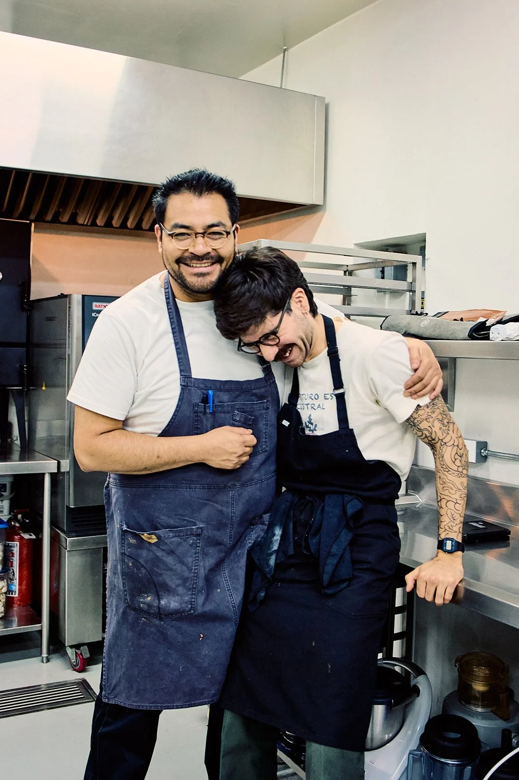 Two men wearing aprons hugging and smiling in a commercial kitchen.