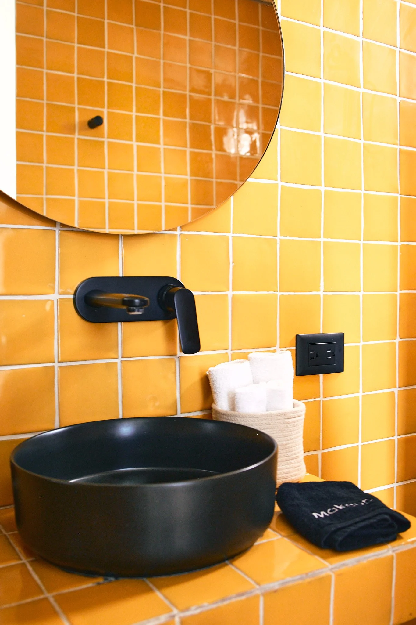 Bathroom with orange tiles, a small round mirror, a black faucet, black bowl, folded white towel, black washcloth, and electrical outlet.