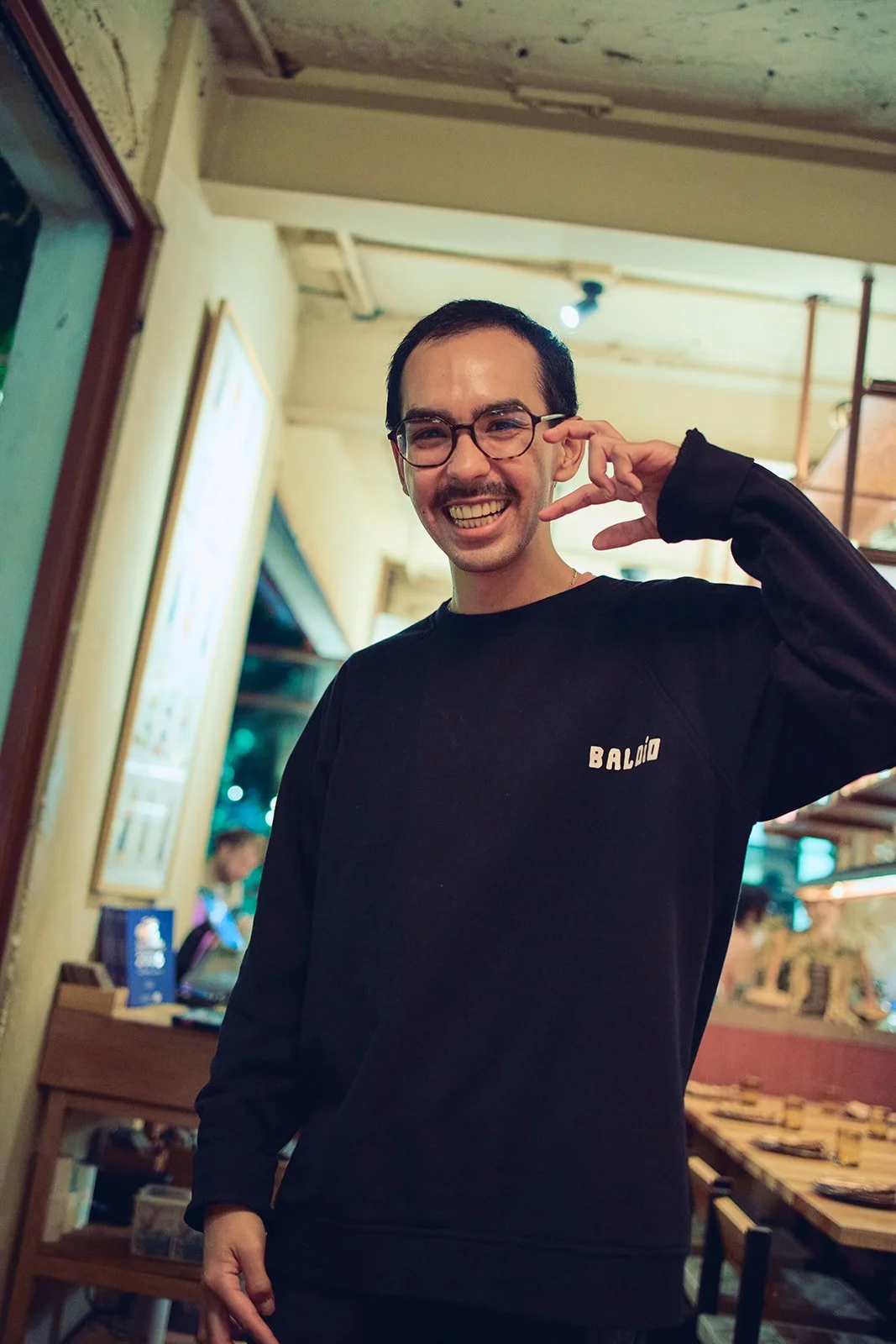Smiling man with glasses in a black Bâldio sweatshirt, making a peace sign gesture in a cozy indoor setting with warm lighting.