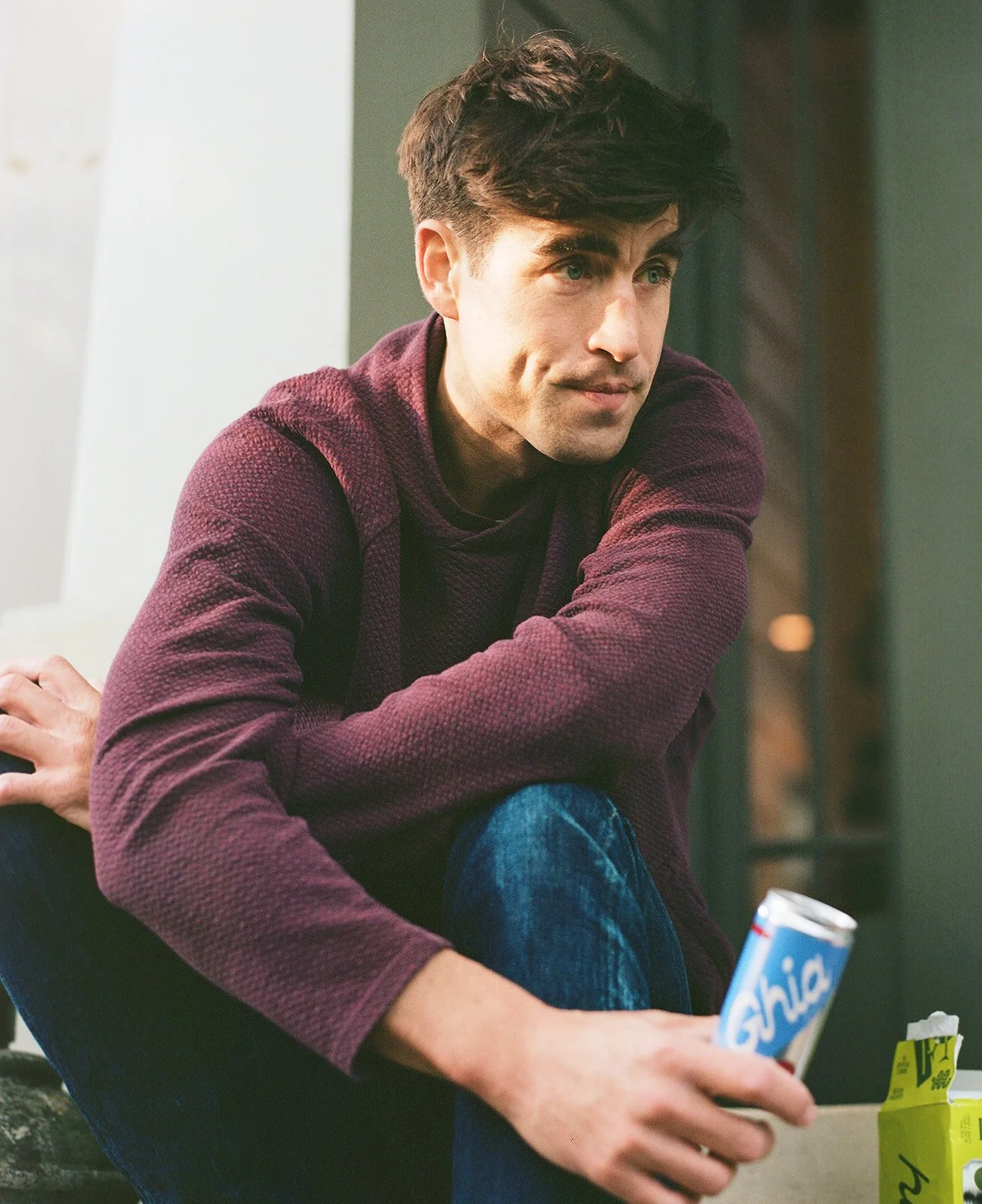 A young man with dark hair and light skin, wearing a maroon textured sweater, sitting on a couch. He is holding an open container of Chips Ahoy cookies in his right hand, with a box of snack or crackers nearby.