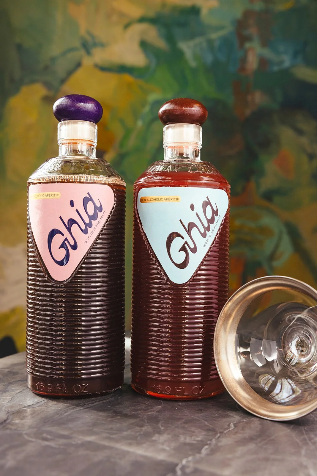 Two bottles of Ghia non-alcoholic aperitif, one with a purple cap and the other with a brown cap, placed on a marble surface against a green and yellow abstract background. One bottle is upright, the other is lying on its side.