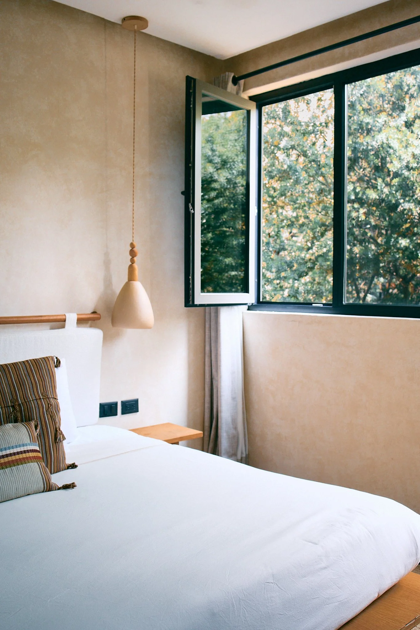 A bedroom with a large bed, a pillow with a striped pillowcase, a wooden bedside table, a hanging light fixture, beige walls, and an open window showing trees outside.