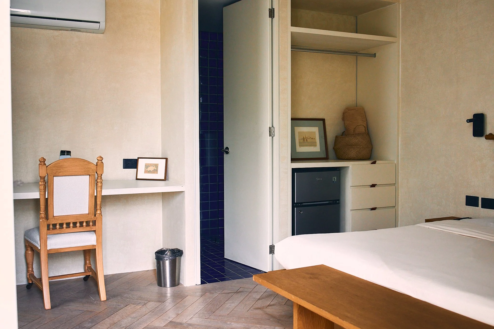 A room with beige walls and white cabinetry, an open closet with a basket and framed picture inside, a mini refrigerator, a wooden table, a wooden chair with a cushioned seat, a small framed picture on the wall, a black electrical outlet, an air cond
