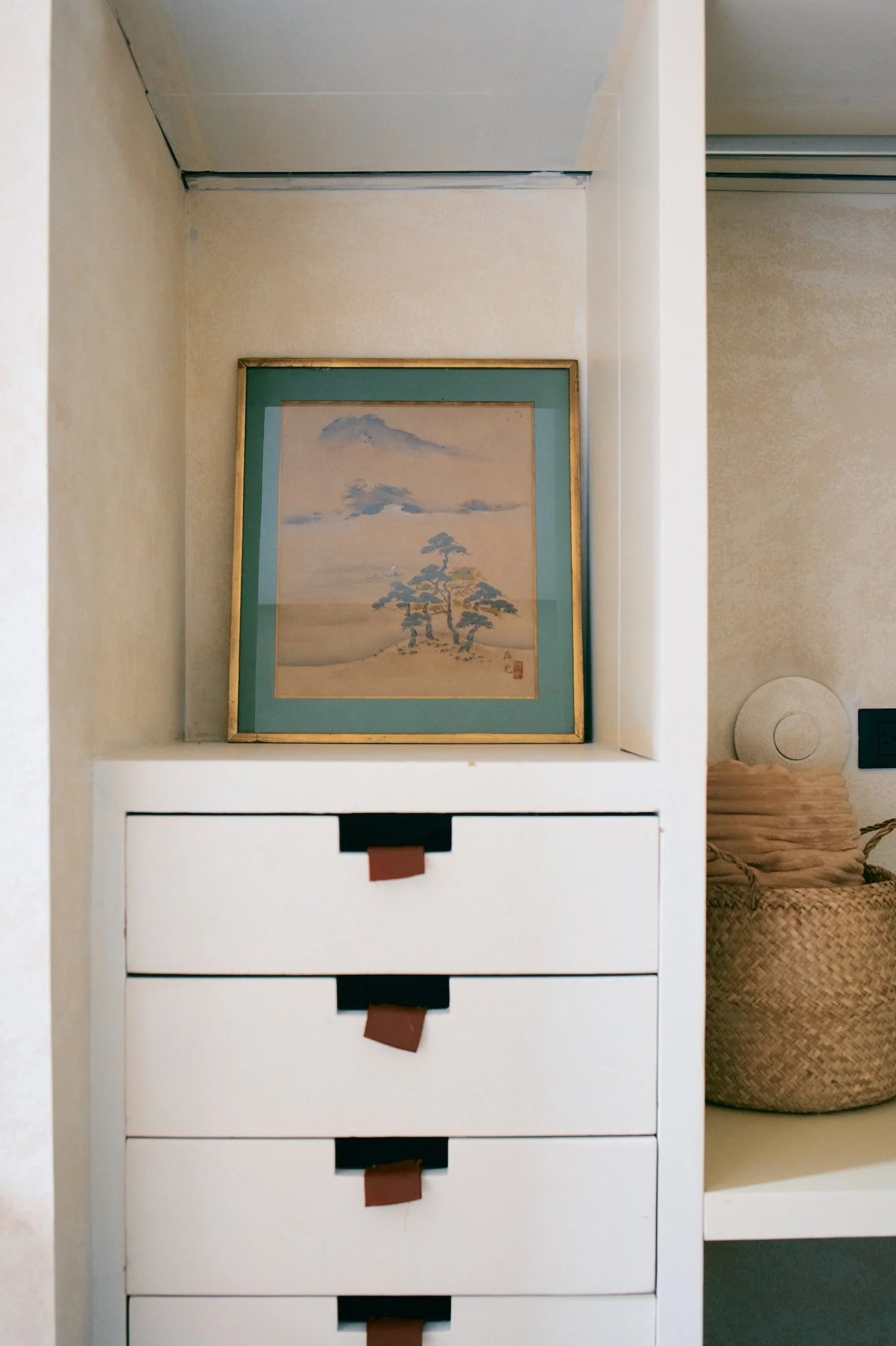 White storage unit with four drawers and brown tabs, positioned in front of a framed painting of a landscape with mountains and trees, with a bathroom or living space visible to the right.