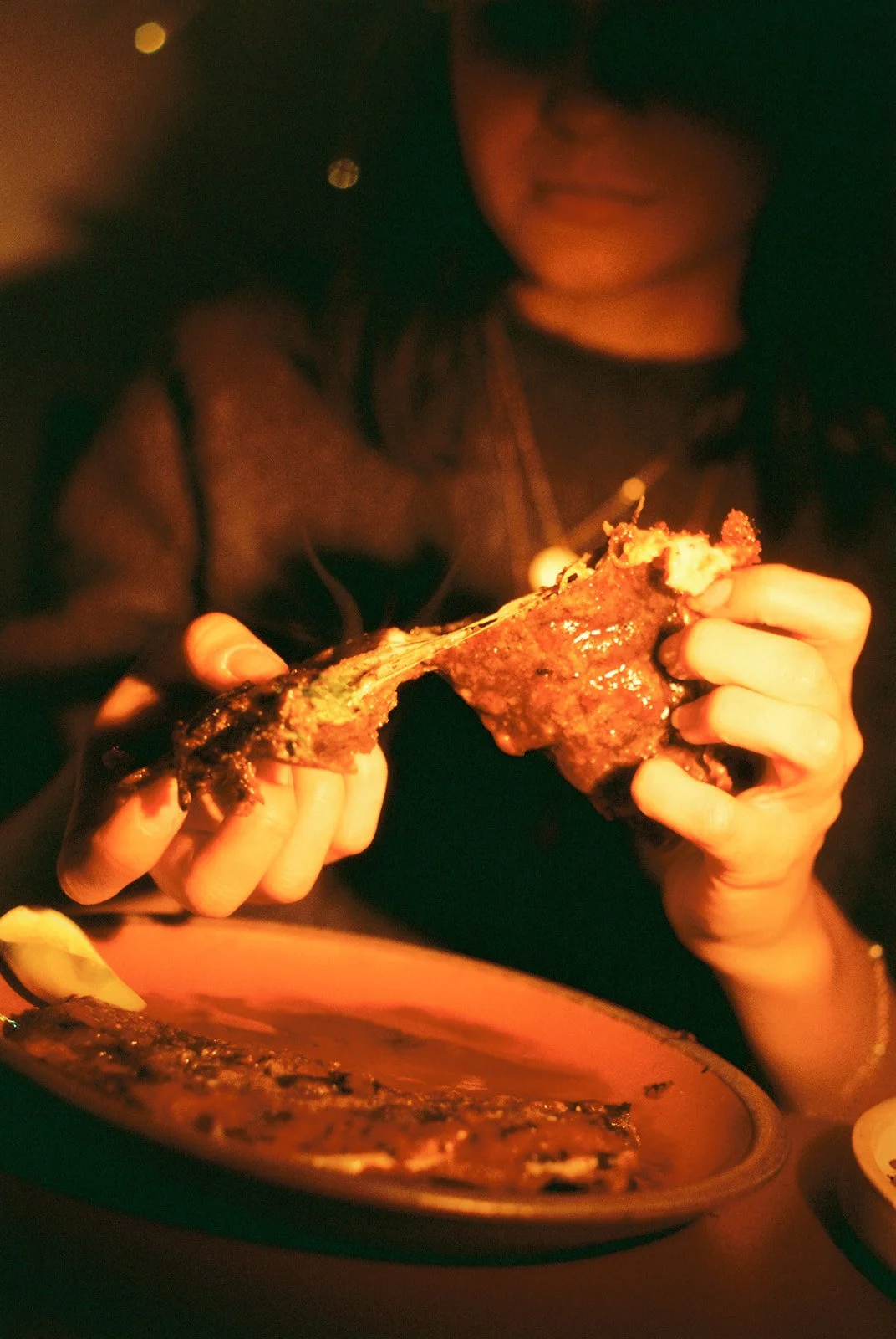 Person holding a piece of fried chicken over a plate of grilled fish in a dimly lit setting.