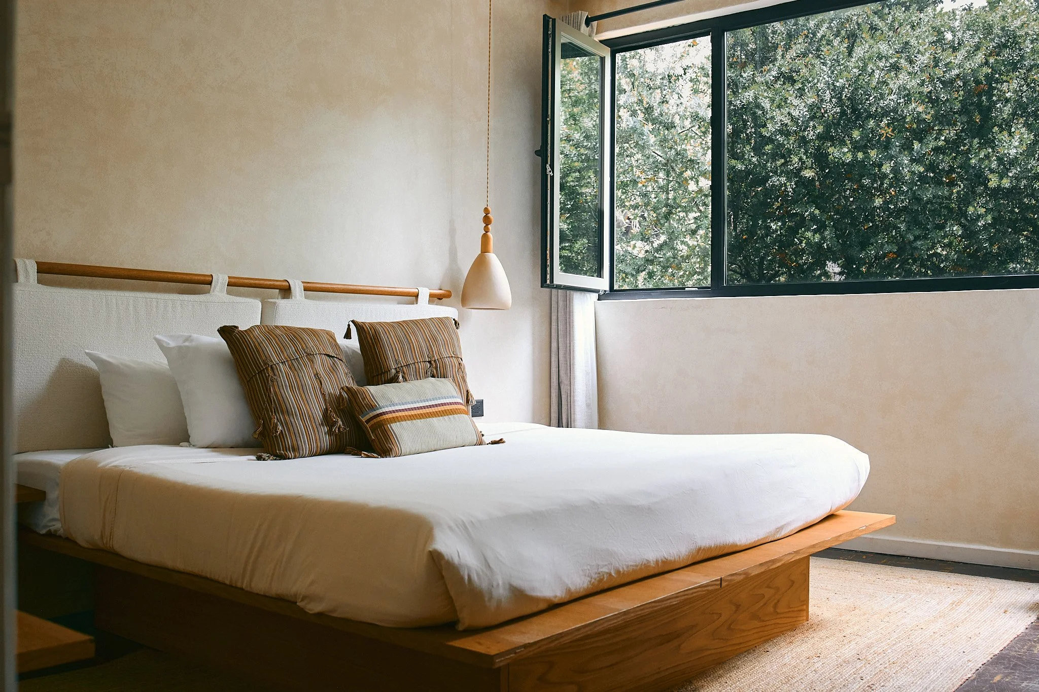 A bedroom with a large bed on a wooden platform. The bed has white linens and several brown and beige striped pillows. There is a beige wall with a hanging pendant light on the left side of the image. An open window with black framing reveals green l
