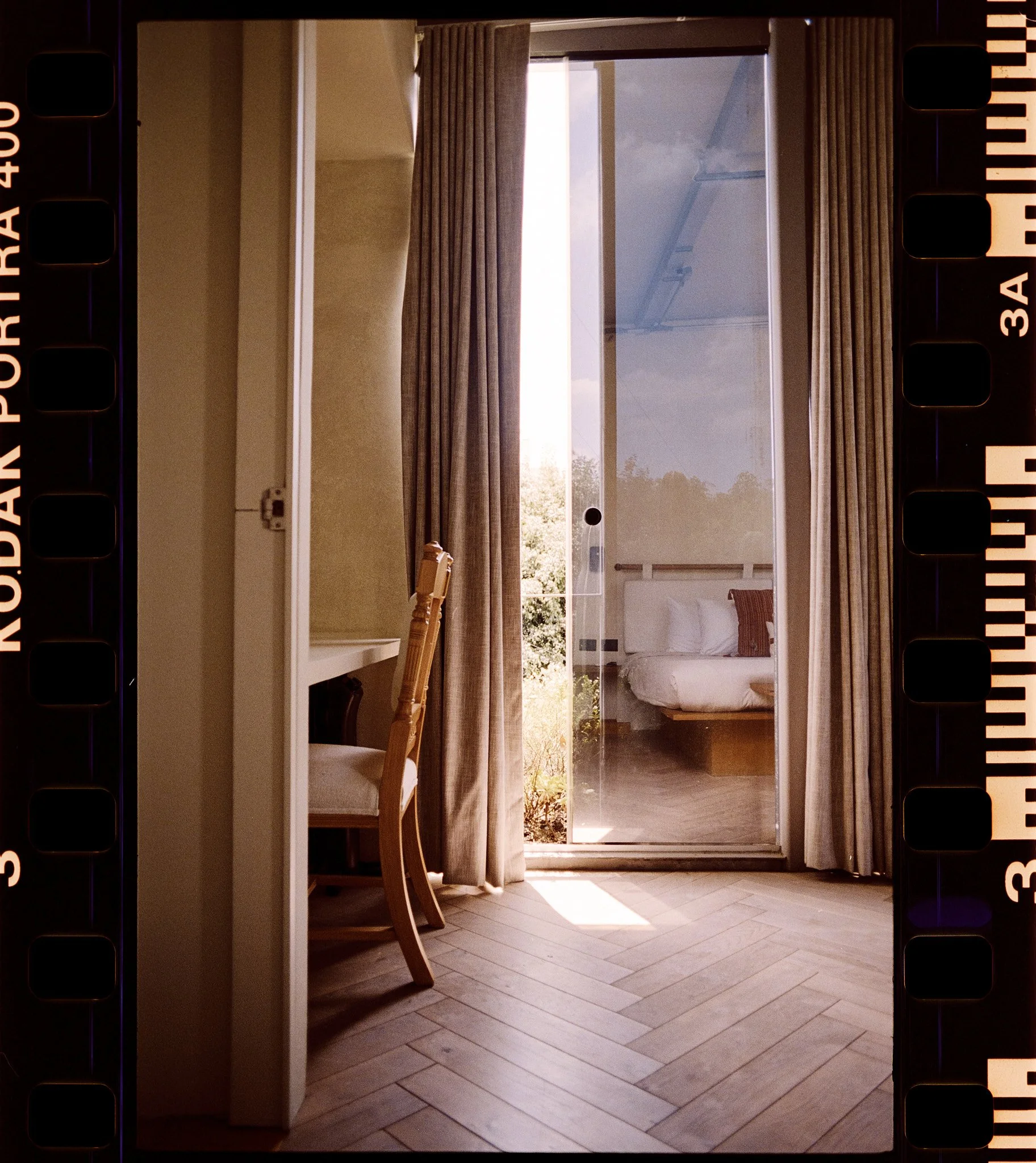 A view from inside a house looking through a sliding glass door to the outside, with beige curtains partially drawn to the sides, a wooden dining chair, and a room with a bed and window outside.