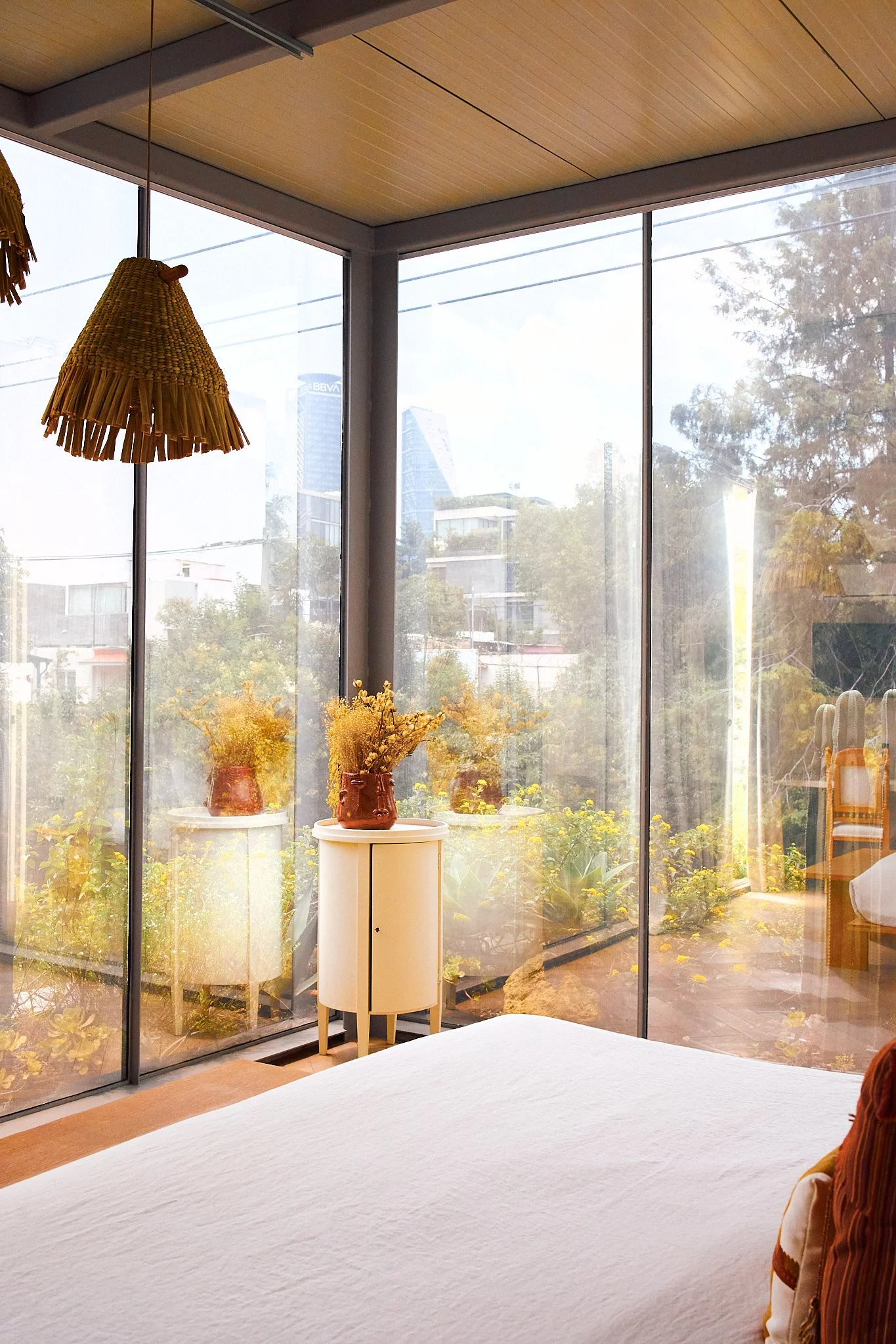 Hotel room with large glass windows showing an outdoor garden and city buildings in the distance. Inside, a bed with white linens is in the foreground, and decorative yellow flowers in vases sit on small tables against the window. There is a hanging 