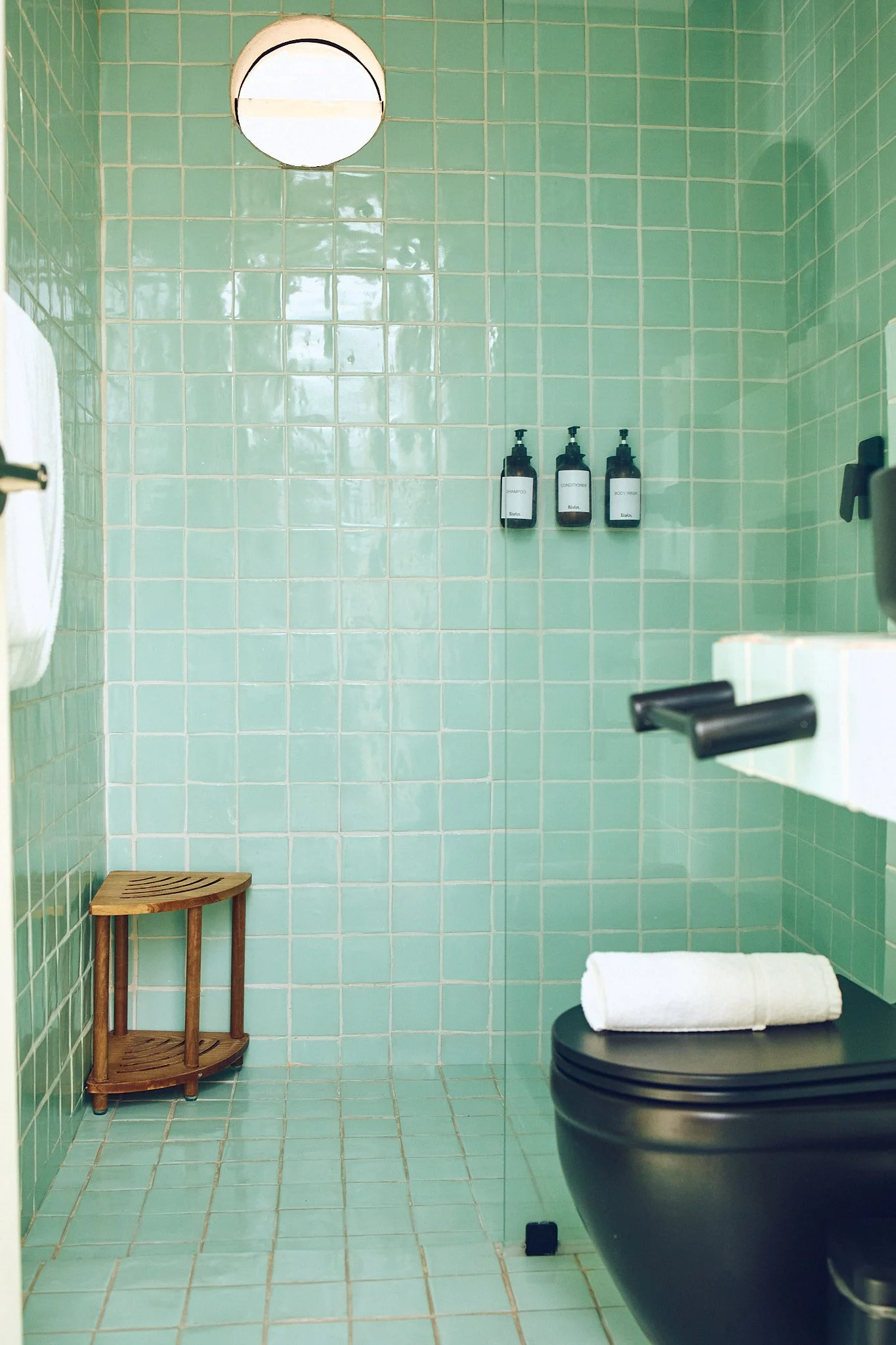 A bathroom with green tiled walls and floor, featuring a wooden stool, a white towel on a black surface, three black bottles labeled 'Shampoo,' 'Conditioner,' and 'Body Wash,' a round mirror, and a black toilet.