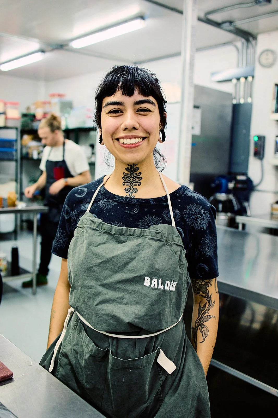 A woman smiling in a kitchen, wearing an apron with the text 'BALD' on it, tattoos on her neck and arms, and dark short hair.