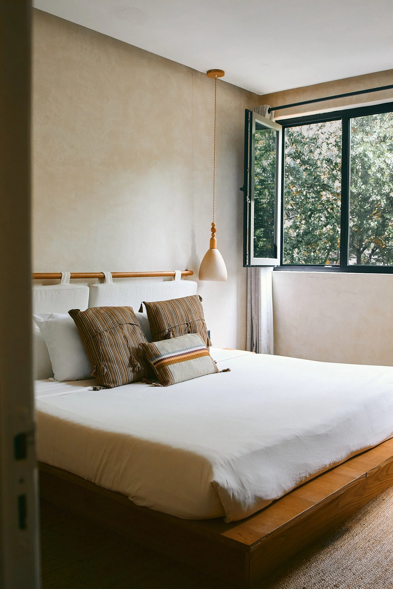 A bedroom with a large bed, striped pillows, a pendant light, and an open window showing trees outside.