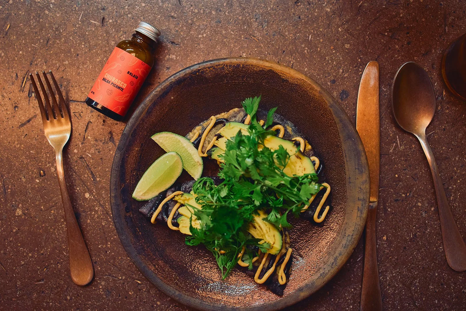A rustic table setting with a bowl of avocado, lime wedges, cilantro, and a drizzle of sauce, accompanied by a fork, knife, spoon, and a bottle of spicy salsa.